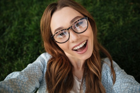 Close up top view of joyful brunette woman in eyeglasses lying on grass in park and making selfieの写真素材