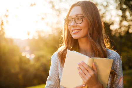 Smiling brunette woman in eyeglasses holding book and looking away in parkの写真素材