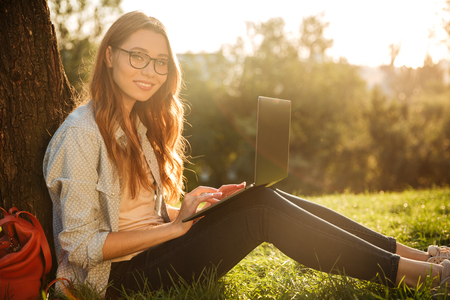 Side view of smiling brunette woman in eyeglasses sitting near the tree in park with laptop computer and looking at the cameraの写真素材