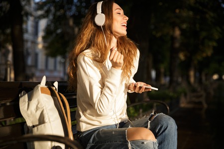 Side view of carefree brunette woman in autumn clothes sitting on bench in park and listening music