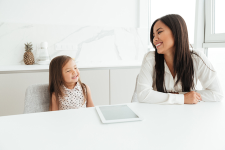 Smiling young asian woman sitting at the kitchen table with her little daughter and using tablet computerの写真素材