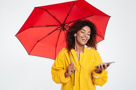 Happy african woman in raincoat hiding under umbrella and listening music on her smartphone over white backgroundの写真素材