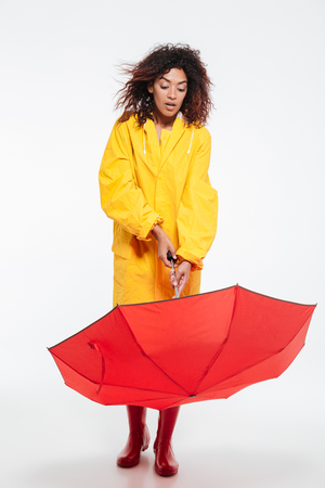 Full length picture of shocked african woman in raincoat opening umbrella over white backgroundの写真素材