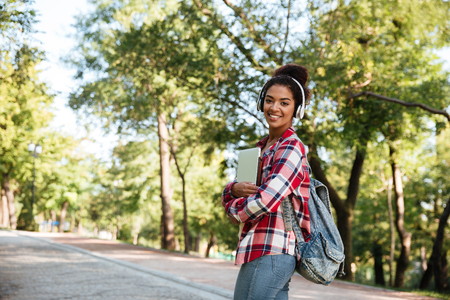 Picture of young african woman walking outdoors in park. Looking aside holding laptop computer listening music by headphones.の写真素材
