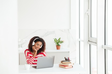 Smiling concentrated african woman using laptop computer by the table near the window