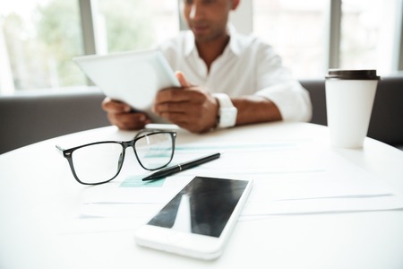 Cropped photo of young african man sitting coworking by tablet computer. Focus on glasses on table.の写真素材
