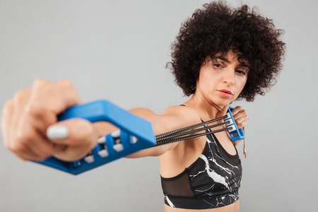 Close up picture of Concentrated curly sports woman doing exercise with hand expander over gray...