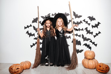 Couple of two little girls dressed in halloween costumes holding brooms and posing with bats and pumpkins on a backgroundの写真素材