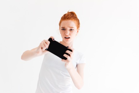 Upset ginger woman in t-shirt playing on her smartphone over white backgroundの写真素材