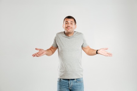 Photo of confused young man dressed in grey t-shirt isolated over grey wall background. Looking...