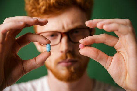Close up portrait of a concentrated redhead man choosing between two pill in his hands isolated over...