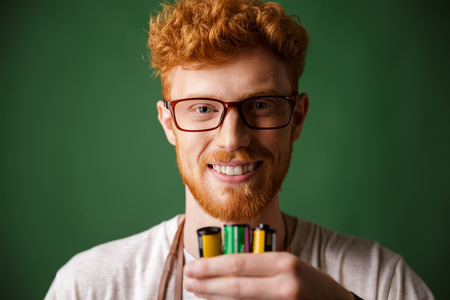 Close-up portrait of readhead bearded photographer in glasses, holding camera rolls, looking at...