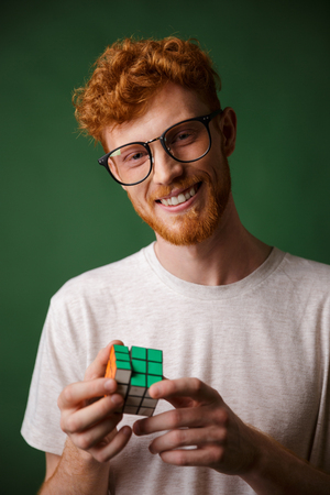 Young smart readgead bearded man in glasses, playing with rubic's cube, looking at camera, over green backgroundの写真素材