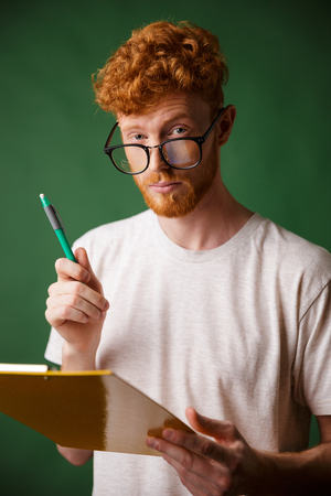 Smart readhead bearded man in white tshirt holding folder and pen, looking at camera, over green backgroundの写真素材