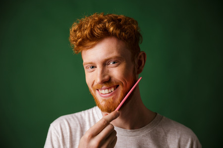 Close-up shot of smiling readhead man, combing his beard with pink comb, over green backgroundの写真素材