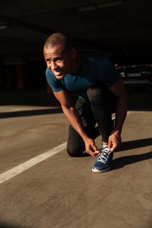 Portrait of a smiling happy afro american sportsman tying his shoelaces while sitting and looking away indoorsの写真素材