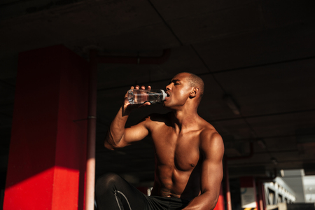 Close up portrait of a young half naked afro american man drinking water after workout indoorsの写真素材