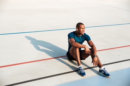 Portrait of a young tired african fitness man in earphones resting while sitting on a ground at the track field outdoors and looking awayの写真素材