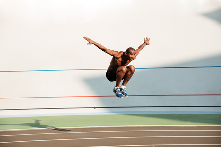 Full length portrait of a half naked fit african sportsman doing a high jump at the track field outdoorsの写真素材
