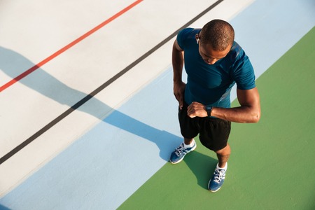 Top view of a young african sportsman looking at his wrist watch while standing on a track field