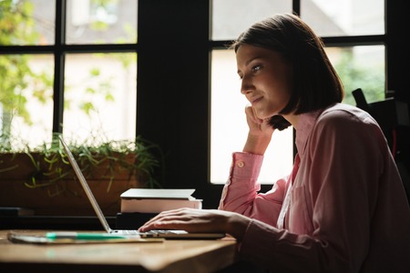 Side view of smiling woman sitting by the table in cafe and using laptop computerの写真素材