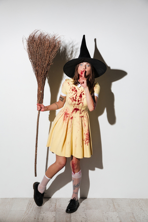 Full length image of young woman in halloween costume posing with broom and looking at the camera while showing silence gesture over white backgroundの写真素材
