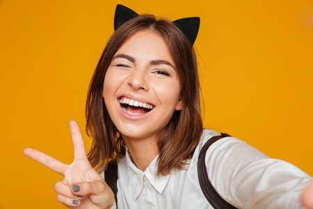 Close up portrait of a happy teenage schoolgirl in uniform with backpack taking a selfie while standing and winking isolated over orange backgroundの写真素材