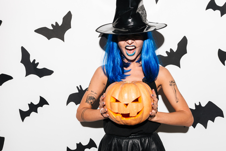 Image of emotional amazing young woman in witch halloween costume wearing hat on party over white background with pumpkin.の写真素材