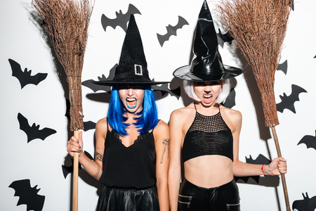 Image of two emotional young women in halloween costumes on party over white background with brooms. Looking camera.の写真素材