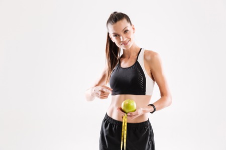 Portrait of a smiling young fitness woman holding green apple and a measuring tape isolated over...