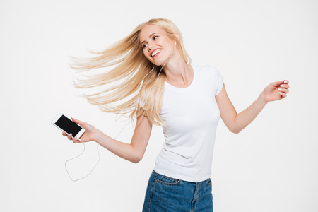 Portrait of a young smiling woman with long blonde hair listening music with earphones and holding mobile phone isolated over white backgroundの写真素材
