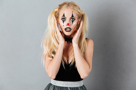 Portrait of a surprised scared blonde woman in halloween clown make-up posing while standing and looking at camera isolated over gray backgroundの写真素材