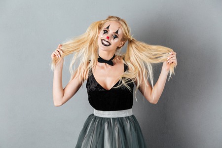 Portrait of a playful blonde woman in halloween clown make-up posing while standing and looking at camera isolated over gray backgroundの写真素材