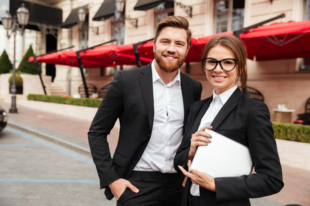 Portrait of a smiling attractive man and woman wearing smart clothes holding tablet computer and looking at camera while standing on a city streetの写真素材