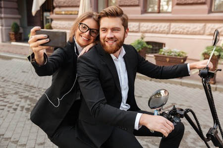 Happy stylish couple satting on modern motorbike outdoors and making selfie on smartphoneの写真素材