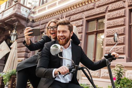 Happy screaming stylish couple satting on modern motorbike outdoors and making selfie on smartphoneの写真素材