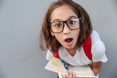 Close up portrait of shocked brunette schoolgirl in eyeglasses holding book and looking at the camera over gray backgroundの写真素材