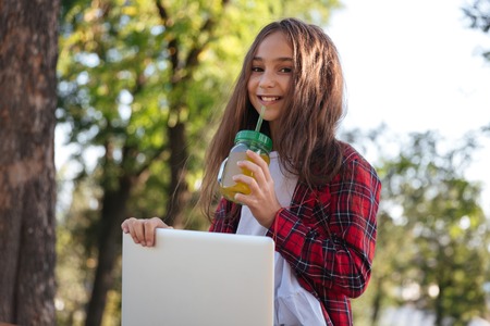 Smiling brunette girl sitting outdoors with laptop computer while drinking juice and looking at the...