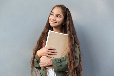 Smiling brunette schoolgirl with long hair dressed in warm clothes holding notebook and looking away over gray backgroundの写真素材