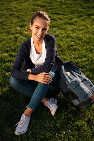 Portrait of a happy cheerful student girl with backpack sitting on the lawn at park and looking at cameraの写真素材