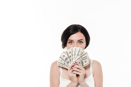 Portrait of an attractive brunette woman covering her face with bunch of money banknotes isolated over white backgroundの写真素材
