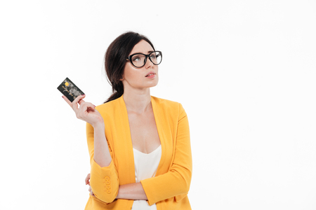 Portrait of an attractive pensive woman in eyeglasses holding credit card and looking away at copy space isolated over white backgroundの写真素材