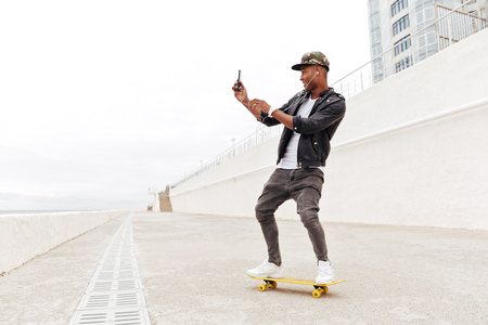 Picture of smiling young african man walking outdoors on skateboard. Looking aside using phone and listening music.の写真素材