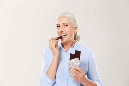 Portrait of cheerful elderly lady eating chocolate, looking at camera, isolated on white backgroundの写真素材