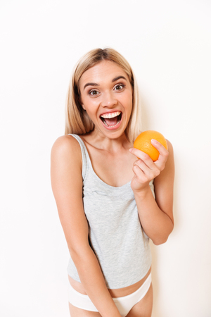 Portrait of a joyful slim girl in underwear holding orange fruit and looking at camera over white wall backgroundの写真素材