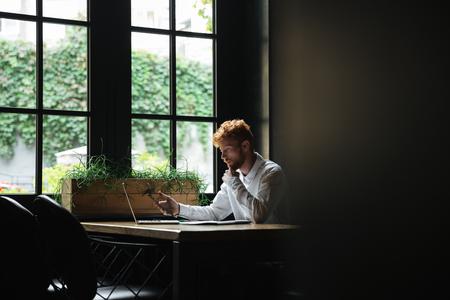 Portrait of serious readhead bearded business man holding smartphone, sitting at workplaceの写真素材