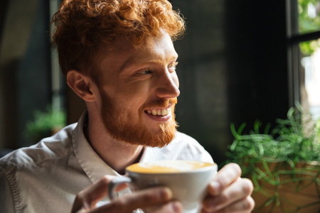 Close-up portrait of handsome smiling readhead bearded man, holding coffee cup, looking asideの写真素材