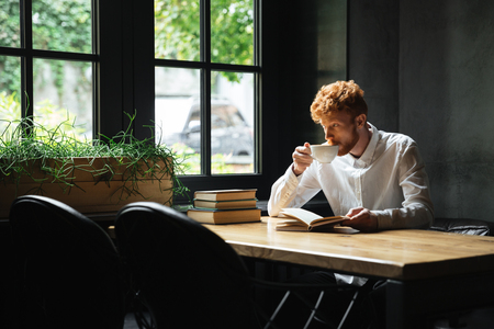Photo of handsome readhead bearded man drinkin coffee in city cafe, looking at large windowの写真素材