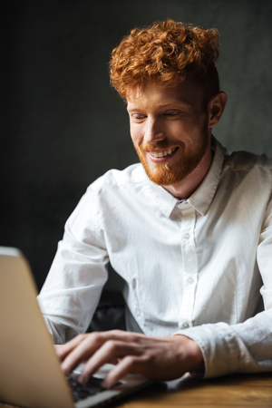 Portrait of a happy young man typing on a laptop while sitting at the table indoorsの写真素材