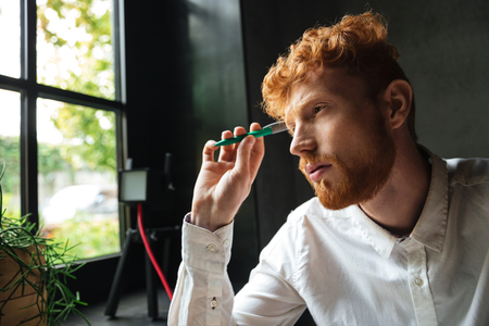 Close-up portrait of young concentrated readhead bearded man, holding green pen, looking at windowの写真素材